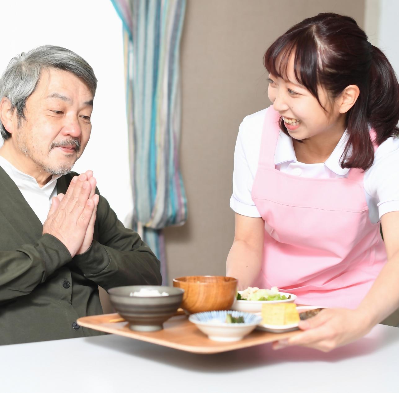 ご飯を運ぶ介護士の女性と、ご飯に手を合わせる高齢の男性