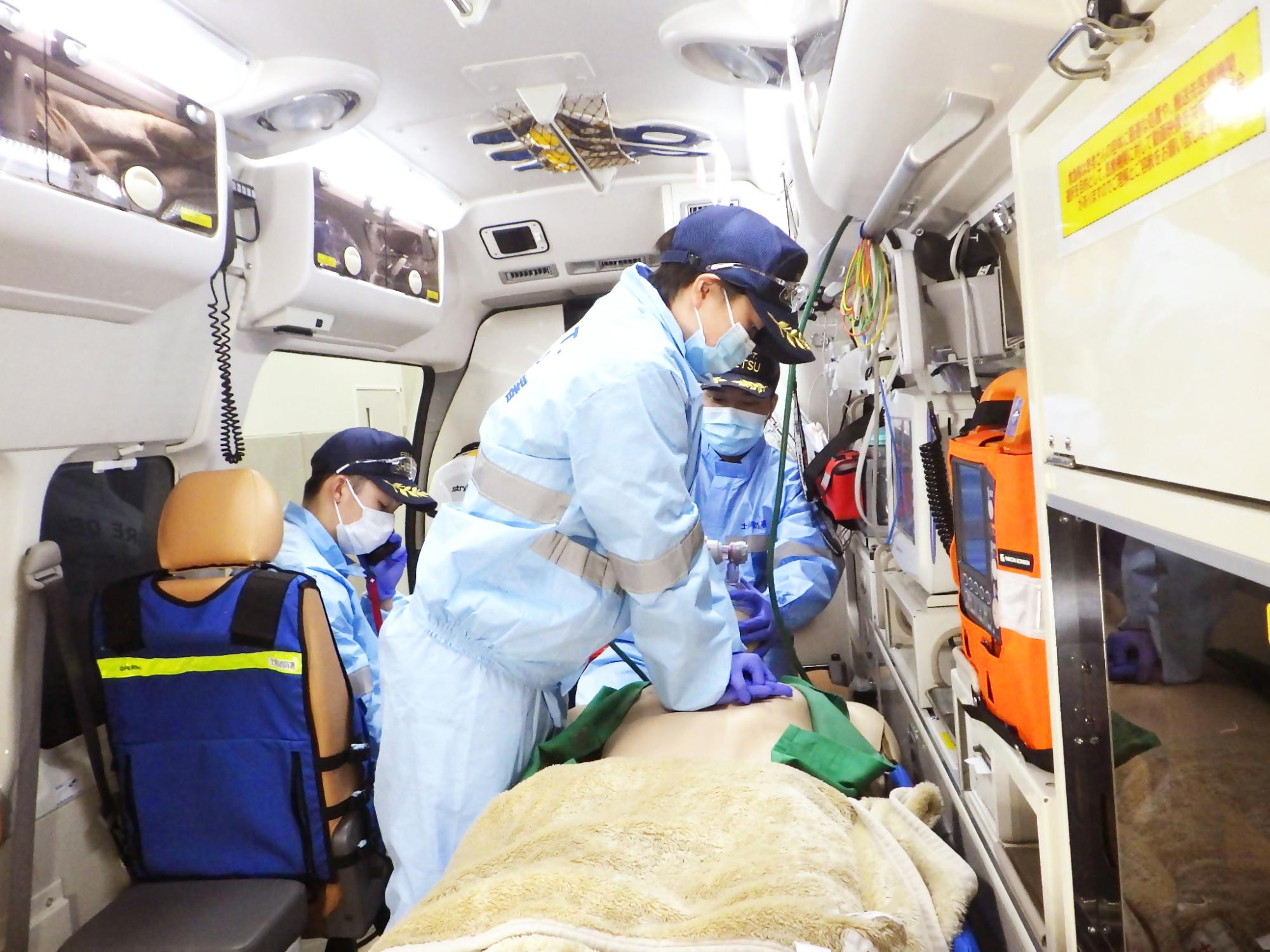 救急救命士の3人が本番を想定した訓練をしている。救急車の中で心肺蘇生や肺に空気を送っていたり、病院に症状を伝えるため電話をしている様子。別の角度から見た写真