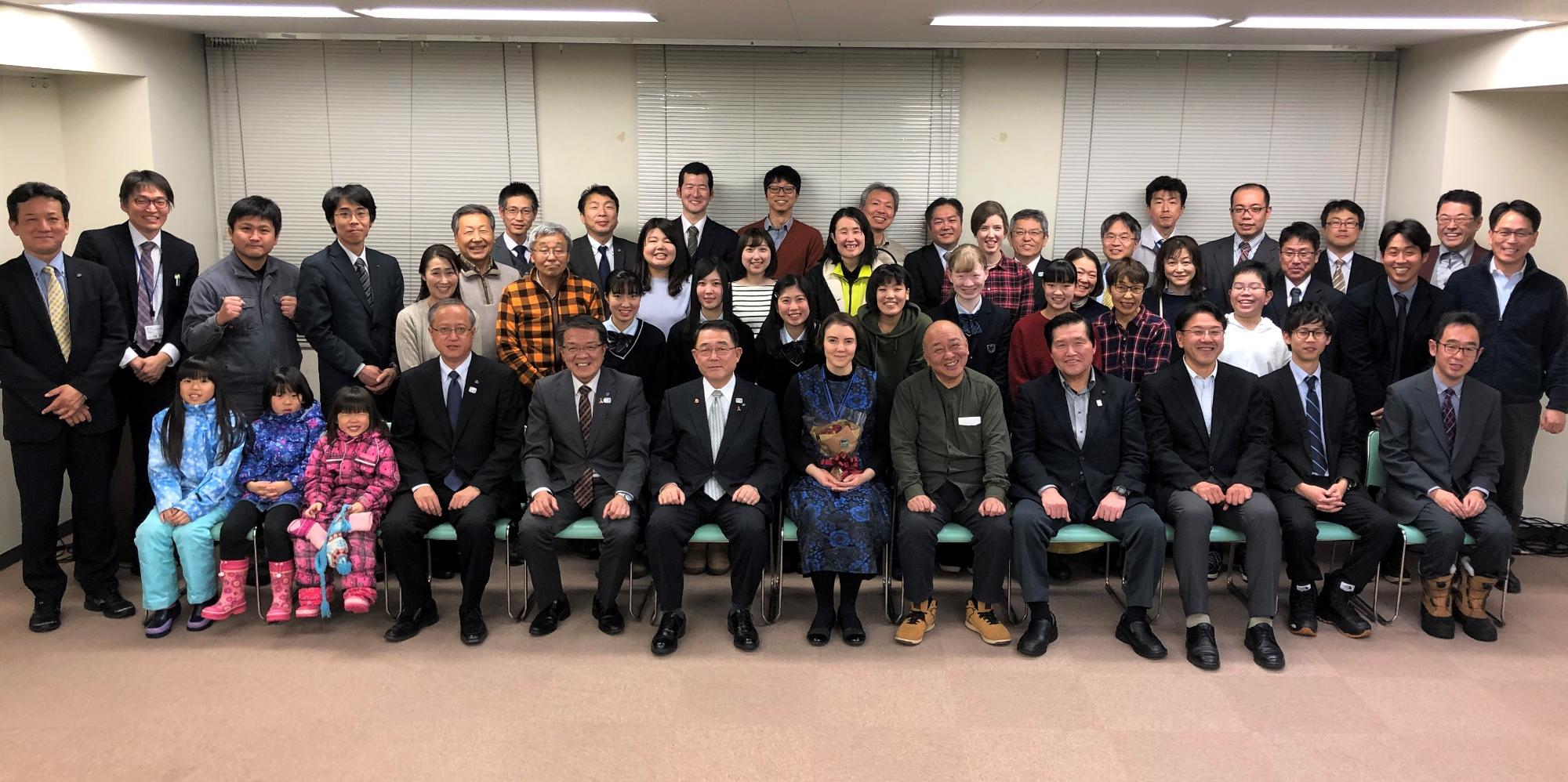 室内の長椅子に座りたくさんの関係者の方々が並んで記念撮影をしている写真