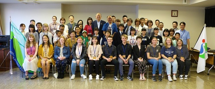 旗が2つ、両端に飾られ、その間に集まり記念撮影をしているたくさんの関係者の人達の写真