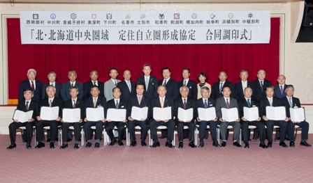北・北海道中央圏定住自立圏構想中心市宣言と書かれた横断幕の下で前列に調印書を持って座っている方々、2列目に立って並んだ記念写真