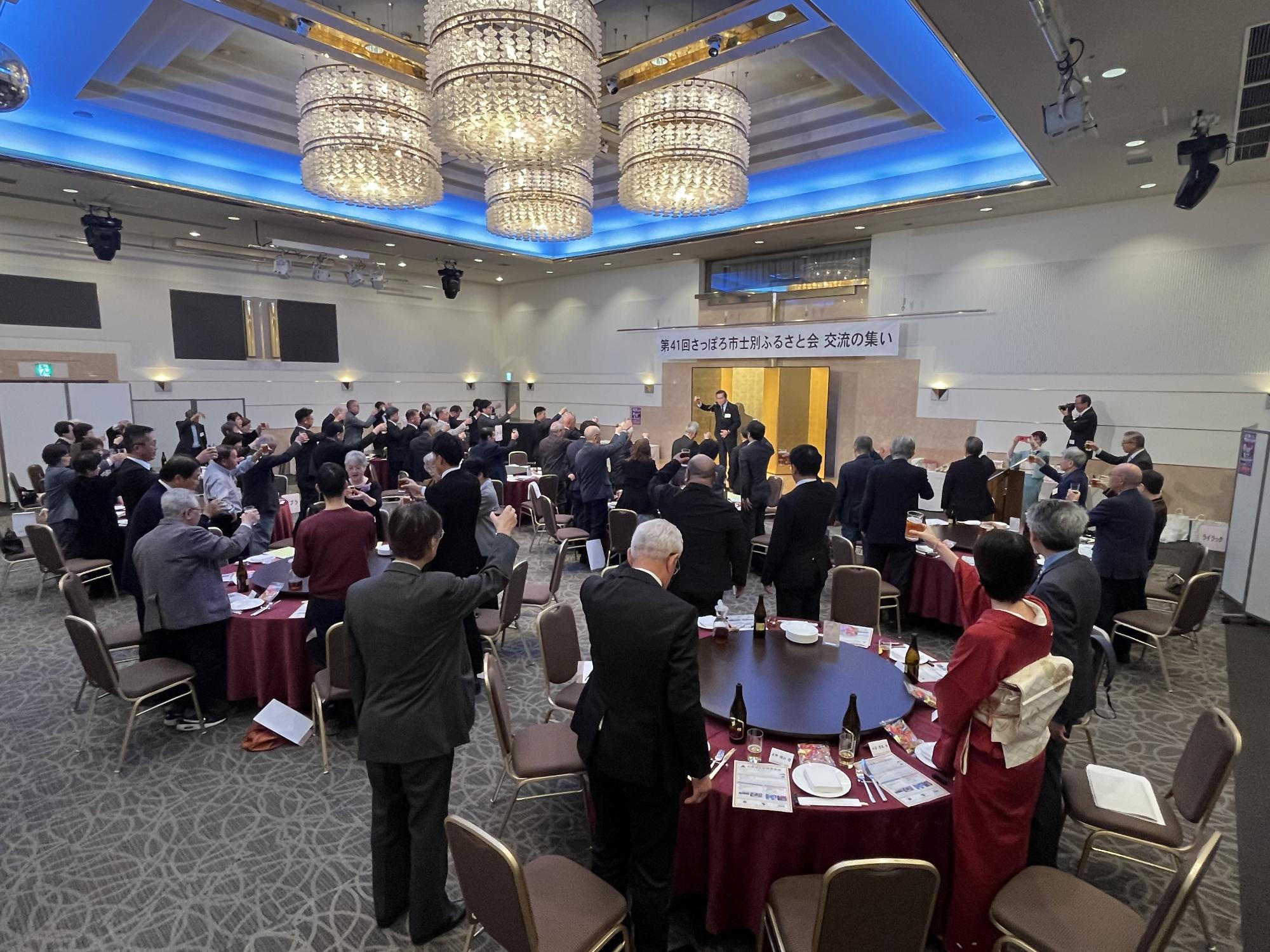 赤いテーブルクロスの丸いテーブルに料理や飲み物が並び、テーブルを囲んで右端にピンクの着物姿の女性がマイクを持ち並び、男性8名女性4名、総勢12名の方が記念撮影をしている写真