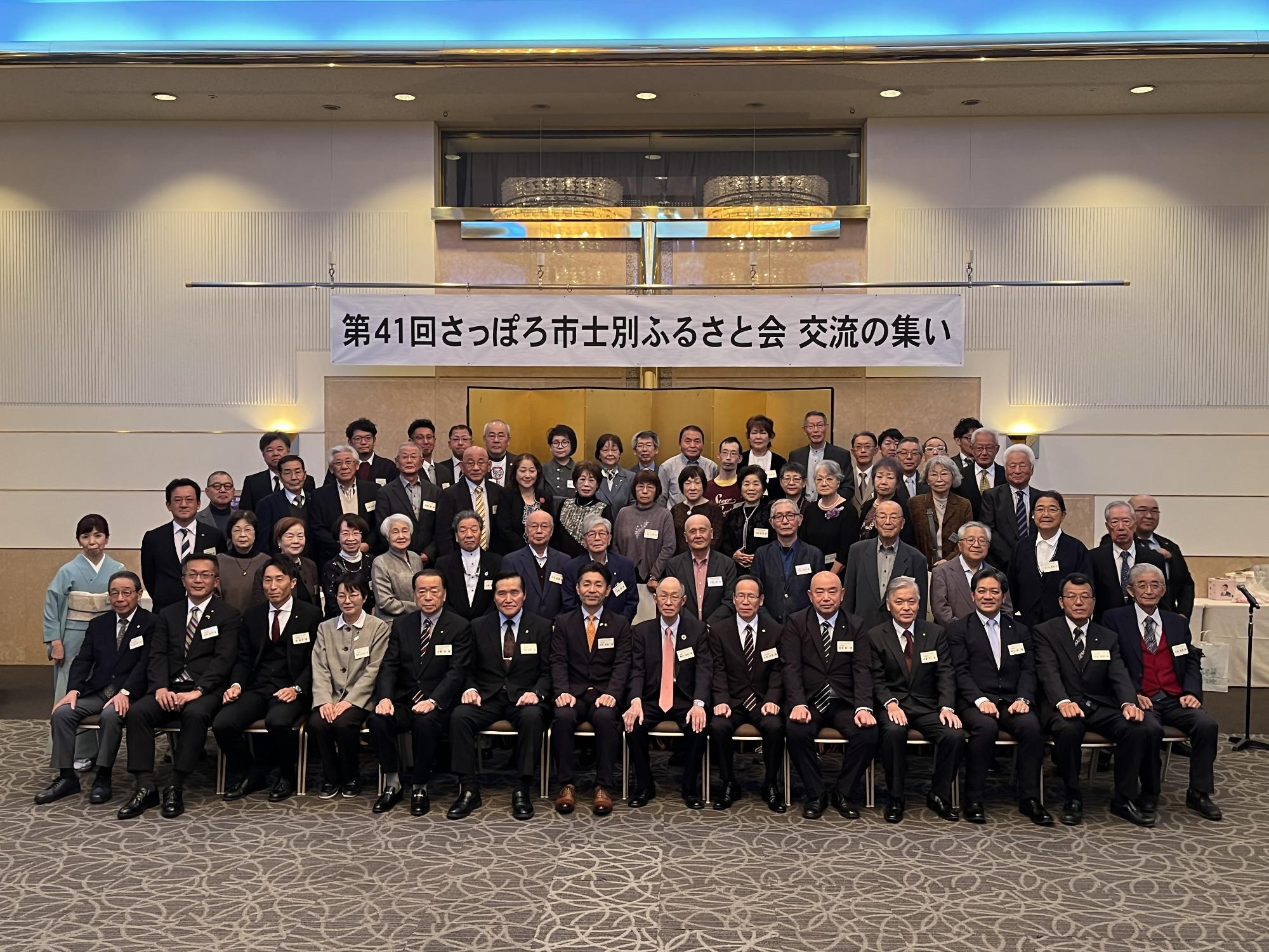 赤いテーブルクロスの丸いテーブルを囲んで右端にピンクの着物姿の女性が並び、男性7名女性5名、総勢12名の方が記念撮影をしている写真