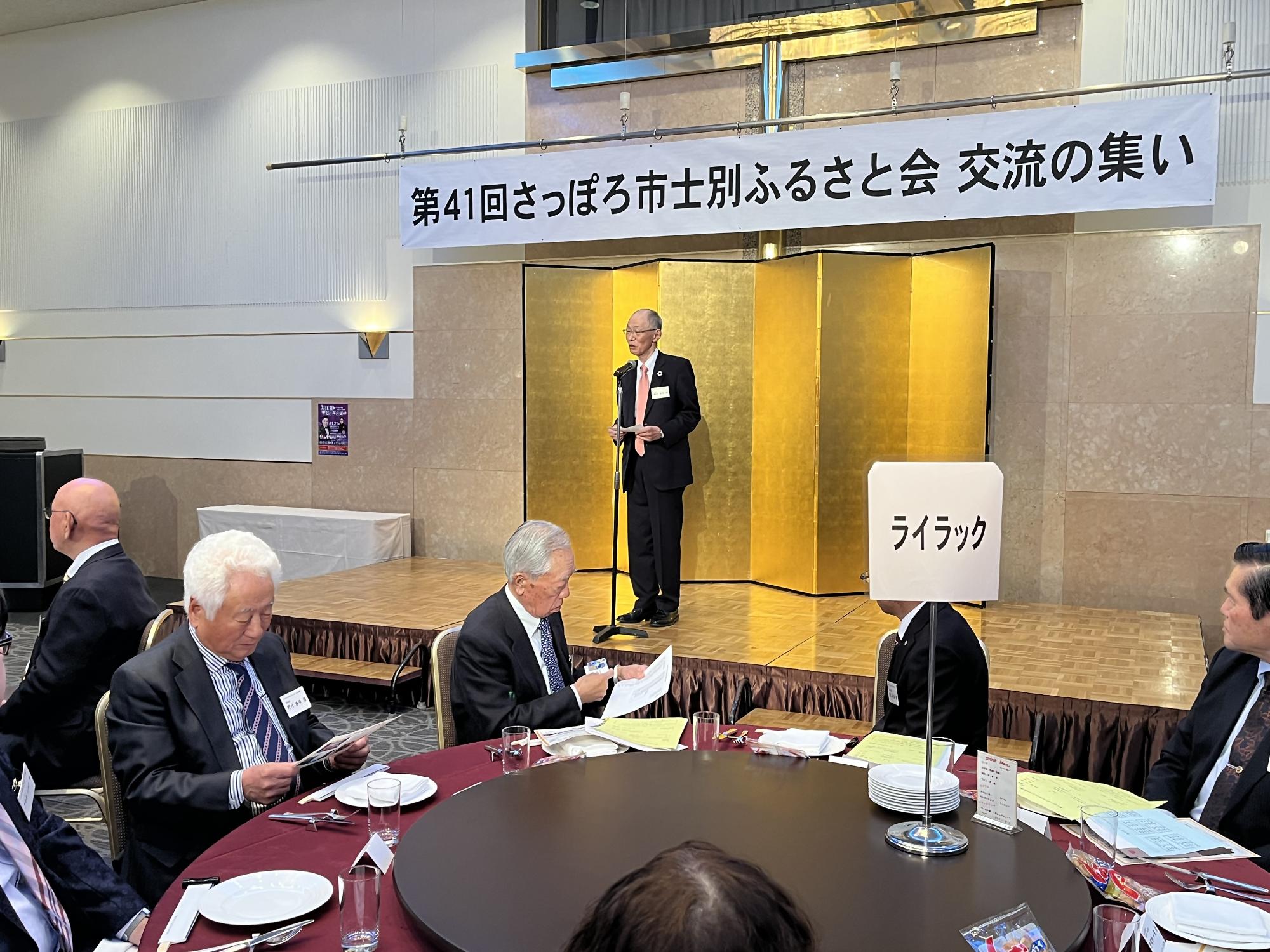 さっぽろ市士別ふるさと会交流の集いの横断幕の下の舞台で1人の男性の挨拶を会場の方々が見ている写真