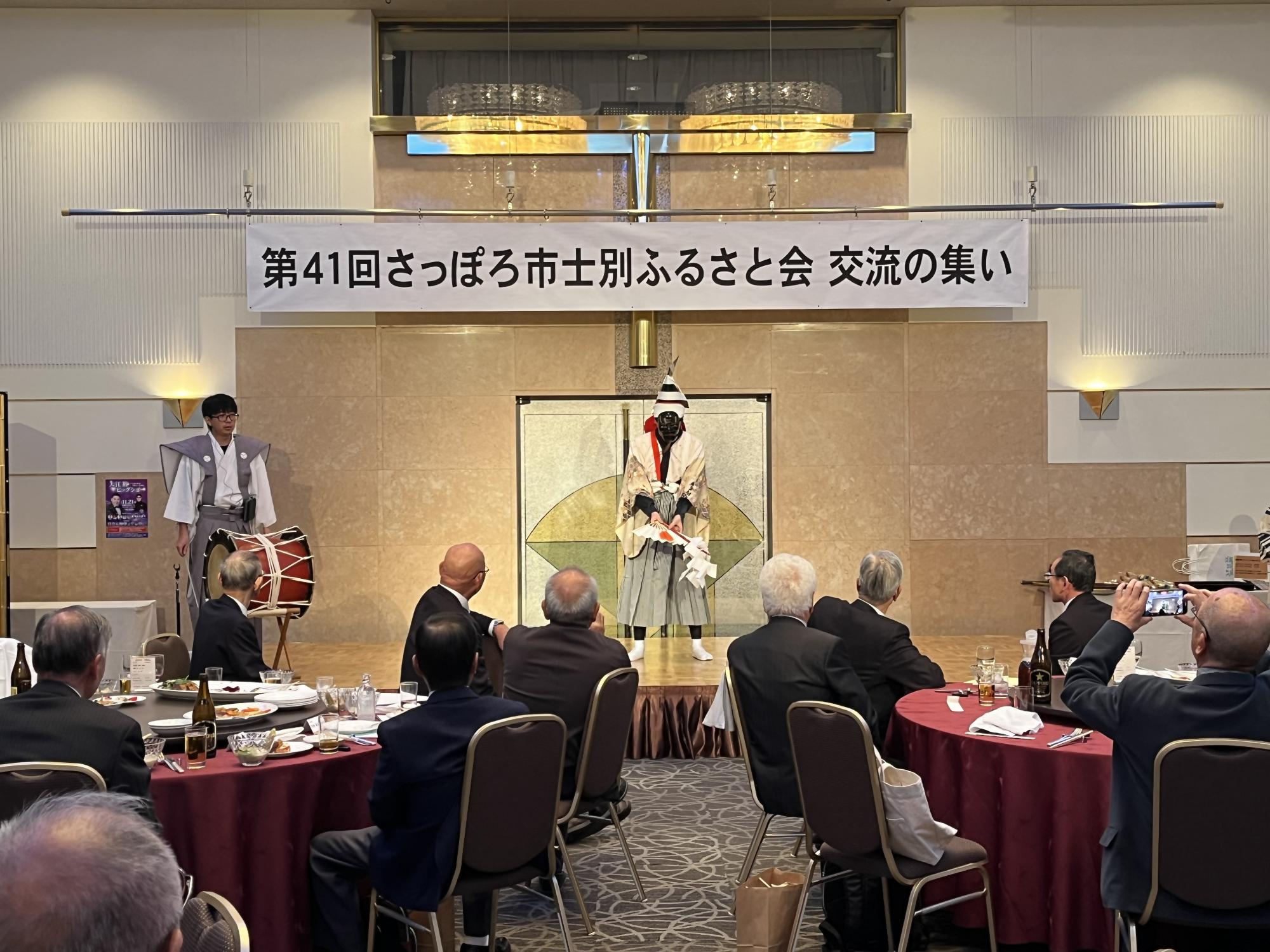 赤いテーブルクロスの丸いテーブルに料理や飲み物が並び、テーブルを囲んで中央にピンクの着物姿の女性が座り、男性11名、総勢12名の方が記念撮影をしている写真