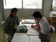 事務所内の長机を挟んで2名の男性が向かい合って座っている土壌診断の様子の写真