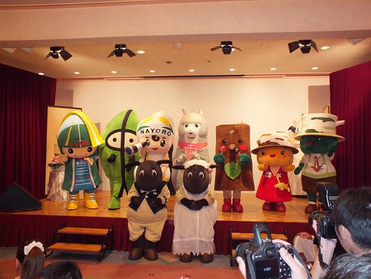 檀上に様々なマスコットキャラクターが並ぶ前でさほっち(羊田黒助くん)とメイちゃん(羊野メイさん)が二人立っており、手前のカメラを持った方々が撮影している様子の写真