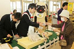 マスコットキャラクターのみーちゃんが見守るなか、制服を着た2名の男子学生が「うんメェ~パイ」の商品を購入に来た女性に販売している様子の写真