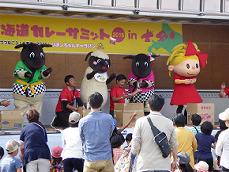 会場のステージ上で4体のマスコットキャラクターが横一列に並んだ間に2名のスタッフが座っているステージイベントの様子の写真