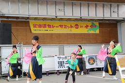 カラフルな衣装を身にまとった4人の女性と1人の子どもがステージ下で踊っている様子の写真