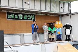 羊まつりのステージ上に「ひつじのショーン」を含めた3体のマスコットキャラクターと男性が右後方に立っている前で、中央のマイクに向かって話をしている女性の写真
