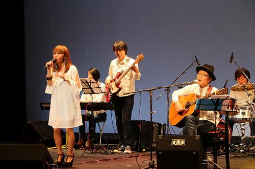 ギターやベースキーボードにドラムを加えた演奏の前でマイクを持って歌っている女性の写真