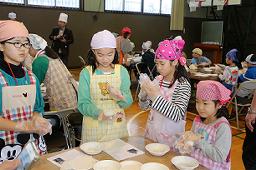 屋内でグループごとに分かれたエプロンと三角巾を着用しビニールの手袋をした4名の女の子がおにぎりを作っている写真