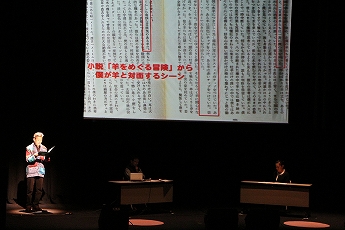 ステージ上に設置された大きなスクリーンに小説が映し出され、左側にスポットライトを浴びている男性がマイクに向かって話をしている様子の写真