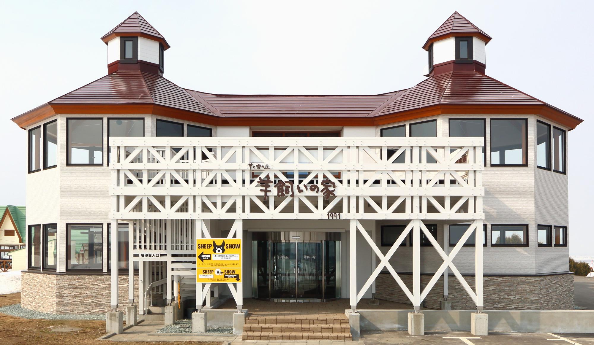 朱色の屋根に白い外壁、窓が沢山あり入り口に黄色い案内看板がたっている羊飼いの家の外観写真