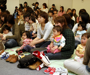 床に敷かれたシートの上に靴を脱いで座り、膝の上に子供を座らせている母親たちの写真
