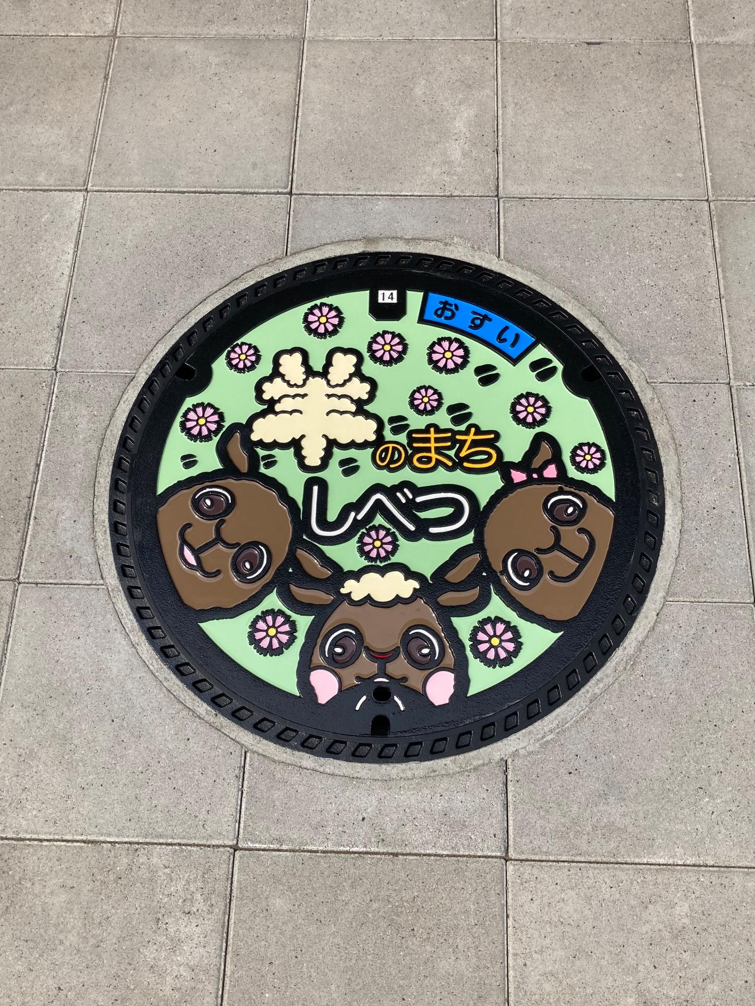 「羊のまちしべつ」の文字にマスコットキャラクター「さほっちファミリー」と散りばめられたコスモスの花が描かれた新しいマンホール蓋のデザインの写真