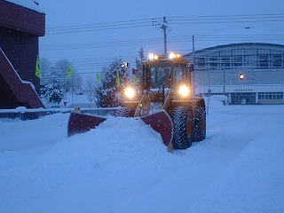 薄暗い中、ライトの付いたホイールローダーで除雪作業を行っている写真