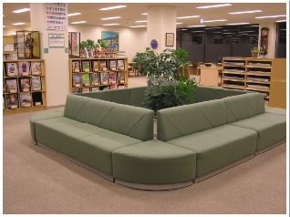 観葉植物を囲むようにカーキー色のソファーが置かれ、本棚には沢山の本が並んでいるブラウジングコーナーの写真