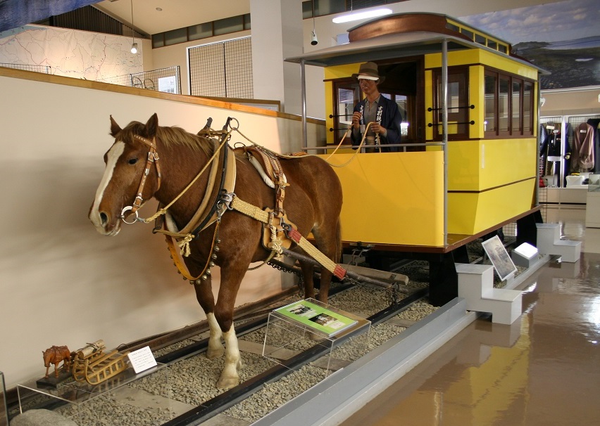 敷かれたレールの上を走る台車に乗る人が手綱を引いて馬を歩かせる様子の人形の写真