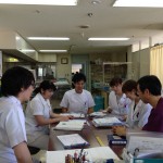 机の上に広げられた栄養管理簿などの資料を見ながら確認し話し合っているNST(栄養サポートチーム)回診の様子の写真