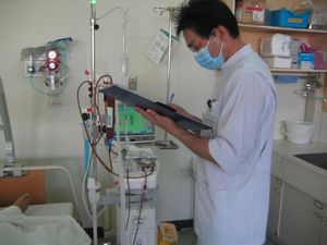 透析の機械の横に立ち、手に持っている資料に記入している白衣を着た臨床工学技士の写真