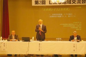 白いあごひげを生やした邉見会長が起立し、両手でマイクを持って話をしている写真