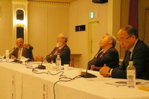 左奥の男性が話をしているのを、両腕を組んで聞いている2名の男性とテーブルの上で両手を組んで話を聞いている男性の写真