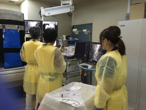 黄色い使い捨てガウンを着た医療従事者3名が画像が映し出されたモニターを見ている写真