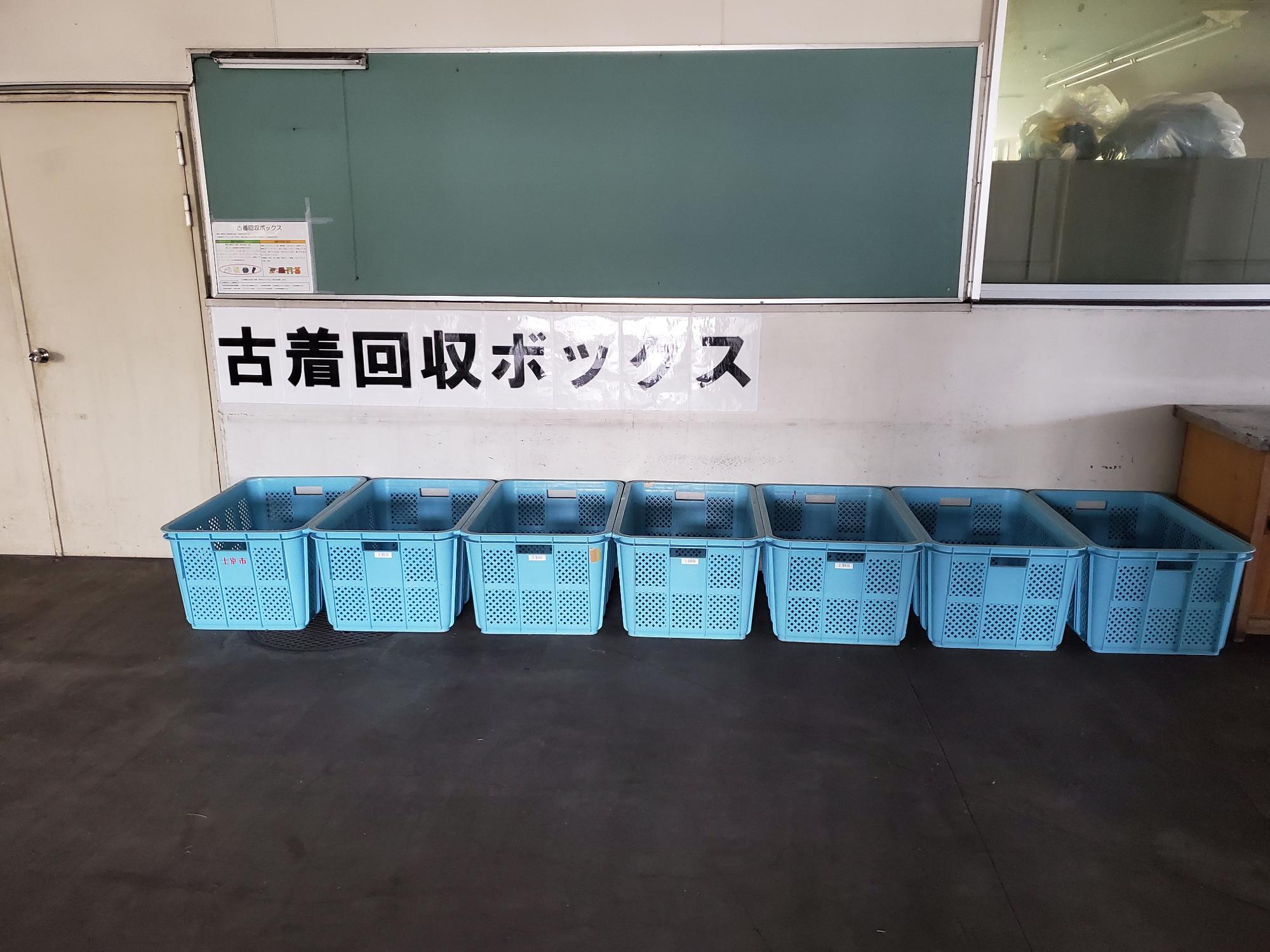 壁に大文字で「古着回収ボックス」と貼られた下に設置された7つの水色のかごが横一列に並んでいる写真