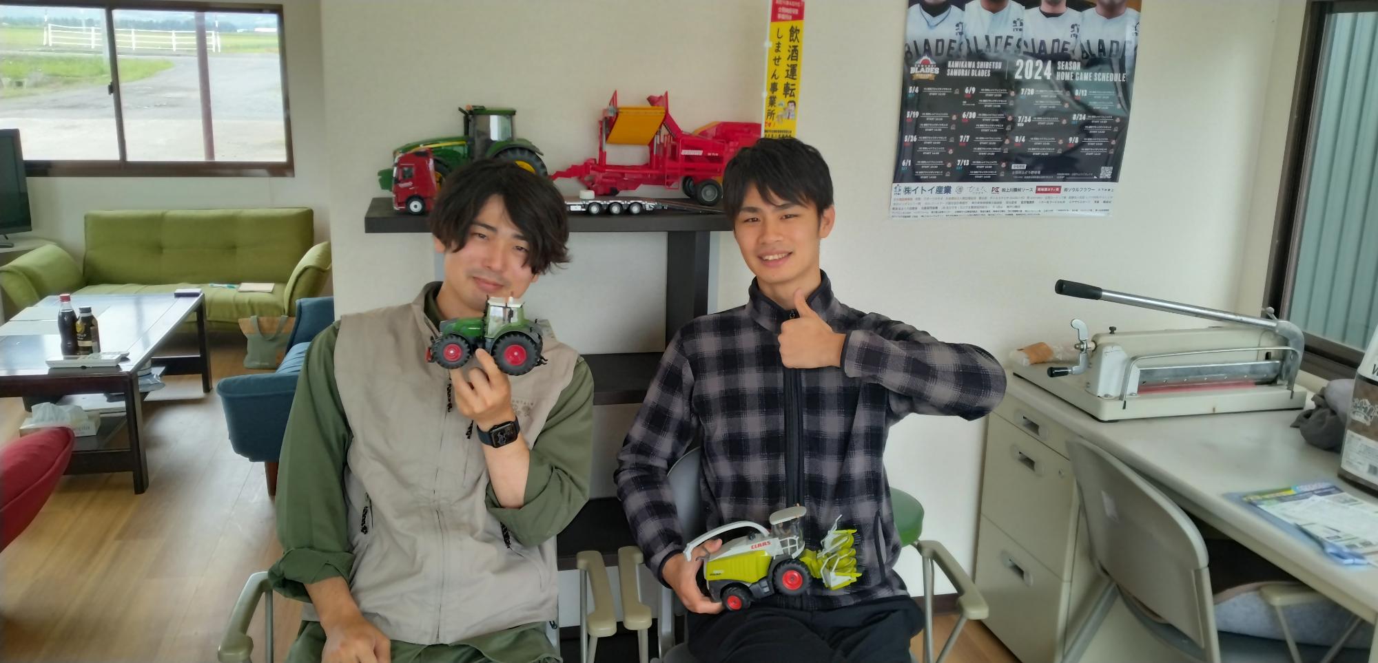緑色のトラクターの模型を左手に持ち微笑んでいる田邉さんと、右手に重機の模型を持ち笑顔で映る木村さんの写真