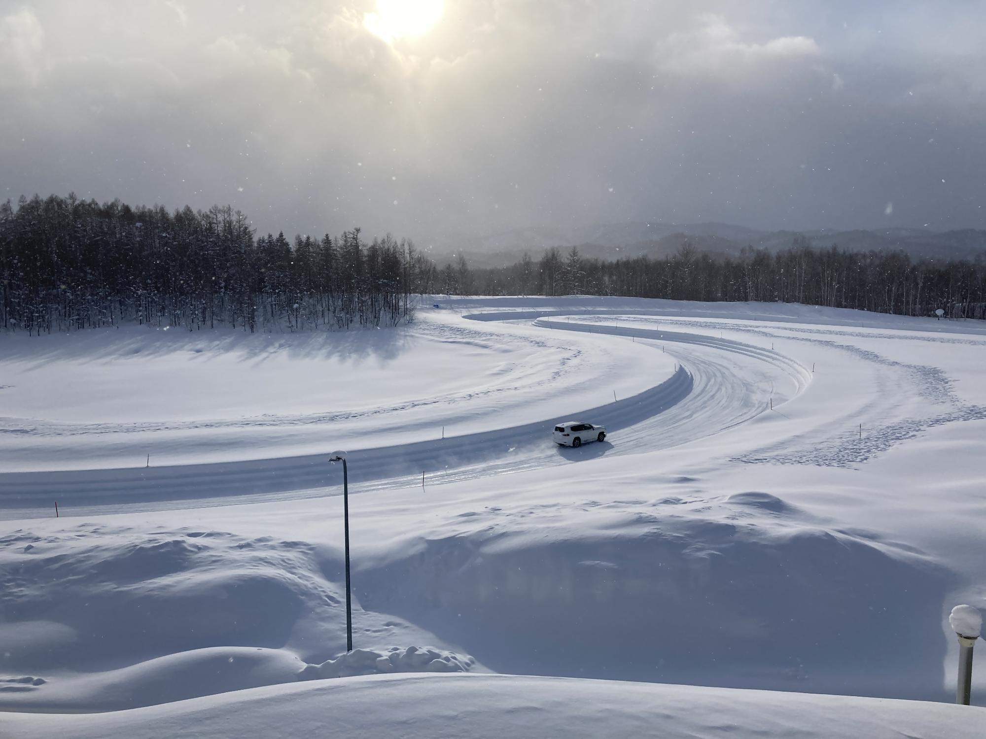 太陽光が降り注ぐ試験場の雪道で自動車走行試験を行っている写真