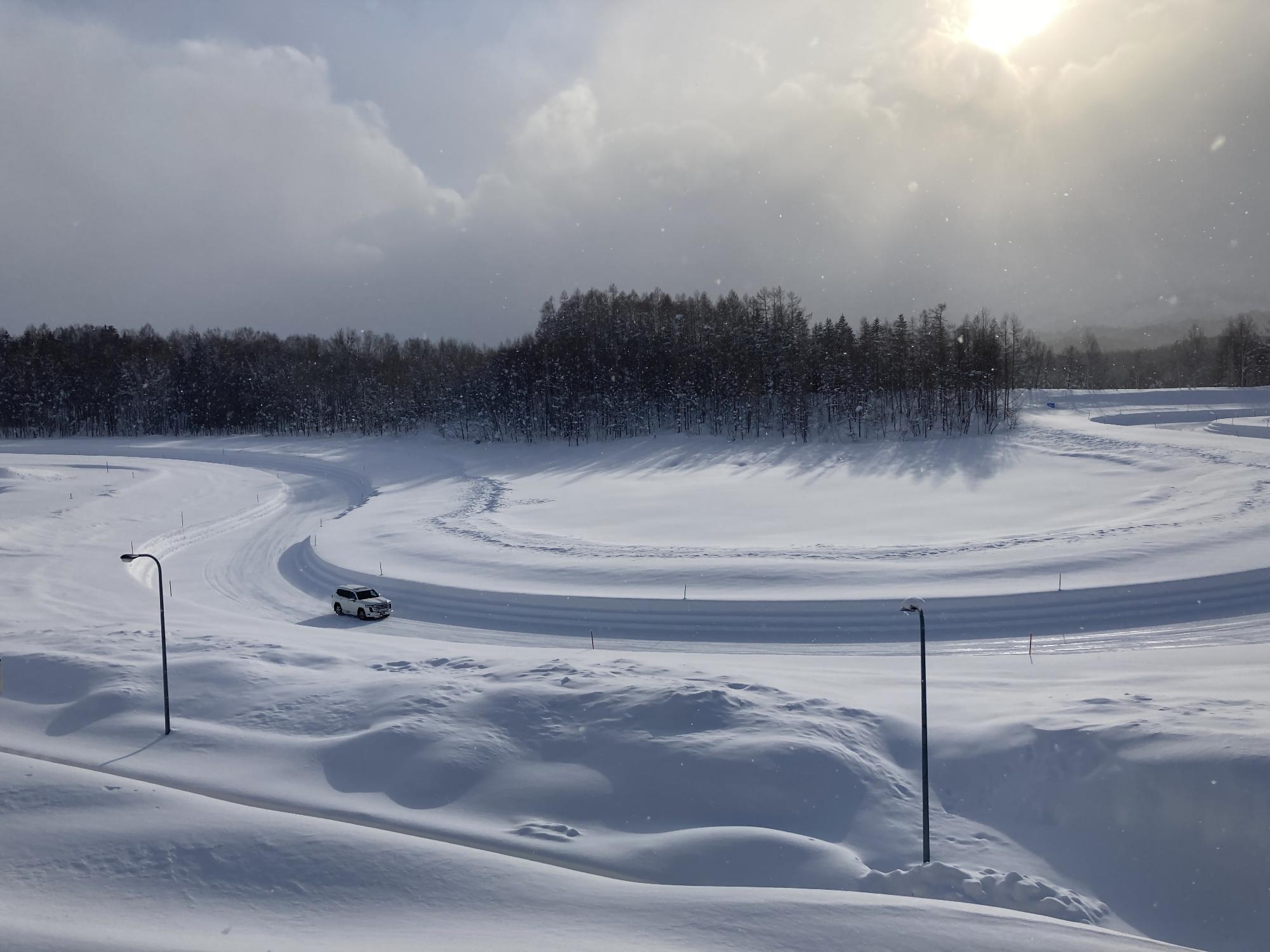 太陽光が降り注ぐ試験場の雪道で自動車走行試験を行っている写真