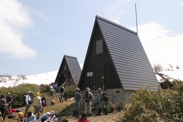 周囲に沢山の登山者が集まっている、縦に細長い三角屋根の2棟の天塩岳避難小屋の写真