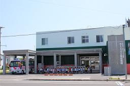 上から白、緑、赤の三色の外壁で園舎の横に園バスが停まっている、認定こども園カトリック士別幼稚園北側の外観写真