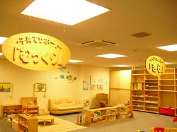 子育てサポート『むっくり』と書かれた木の看板が天井から吊り下げられており、おもちゃや本棚、ソファなどが置かれている室内の写真