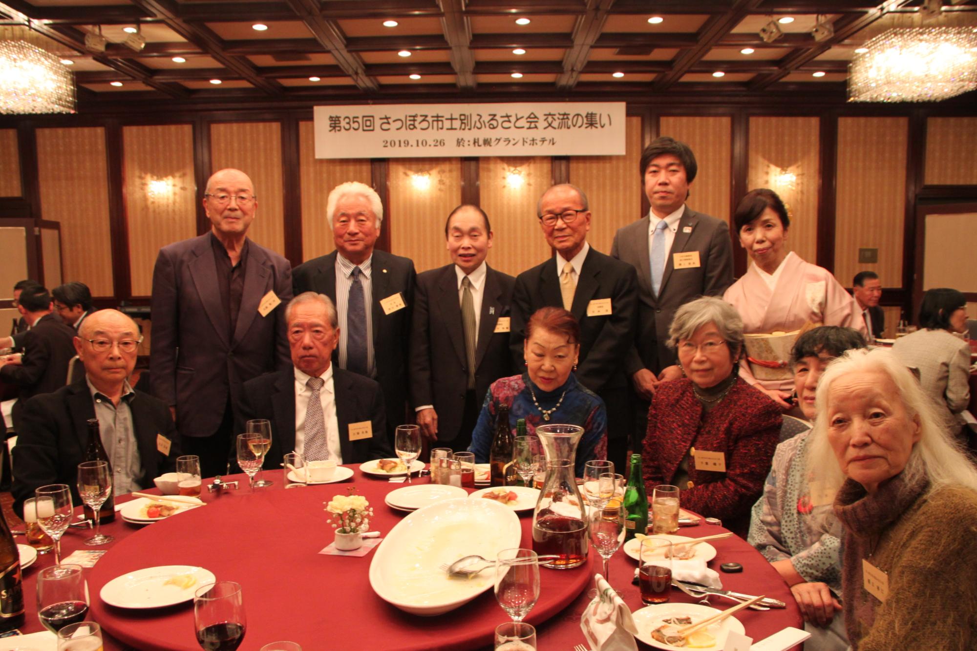 赤いテーブルクロスの丸いテーブルに料理や飲み物が並び、テーブルを囲んで右端にピンクの着物姿の女性が並び、男性7名女性5名、総勢12名の方が記念撮影をしている写真