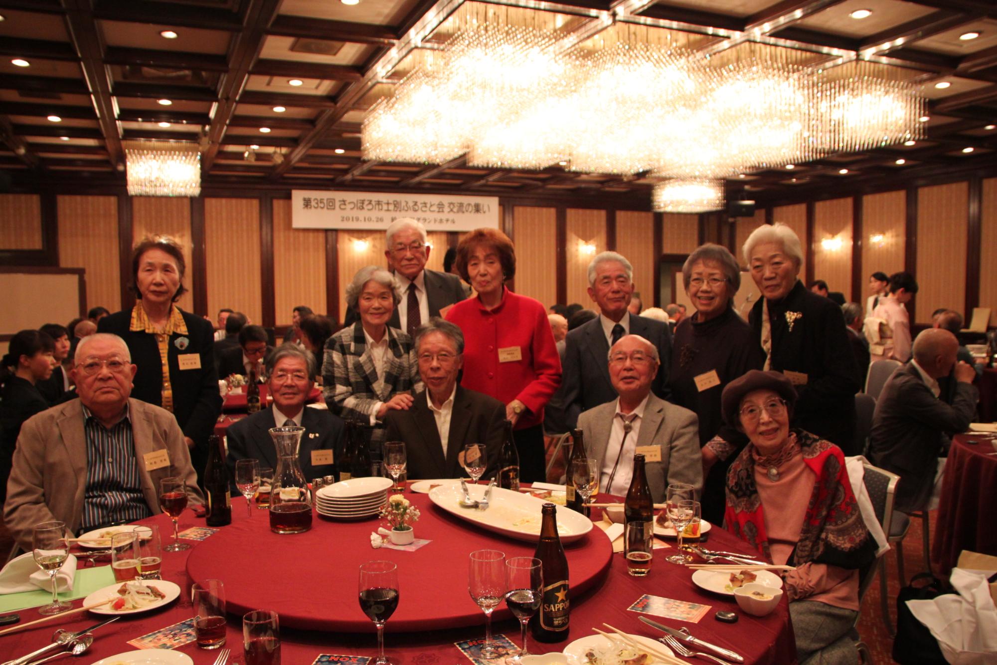 赤いテーブルクロスの丸いテーブルに料理や飲み物が並びテーブルを囲んで、男性7名女性5名、総勢12名の方が記念撮影をしている写真