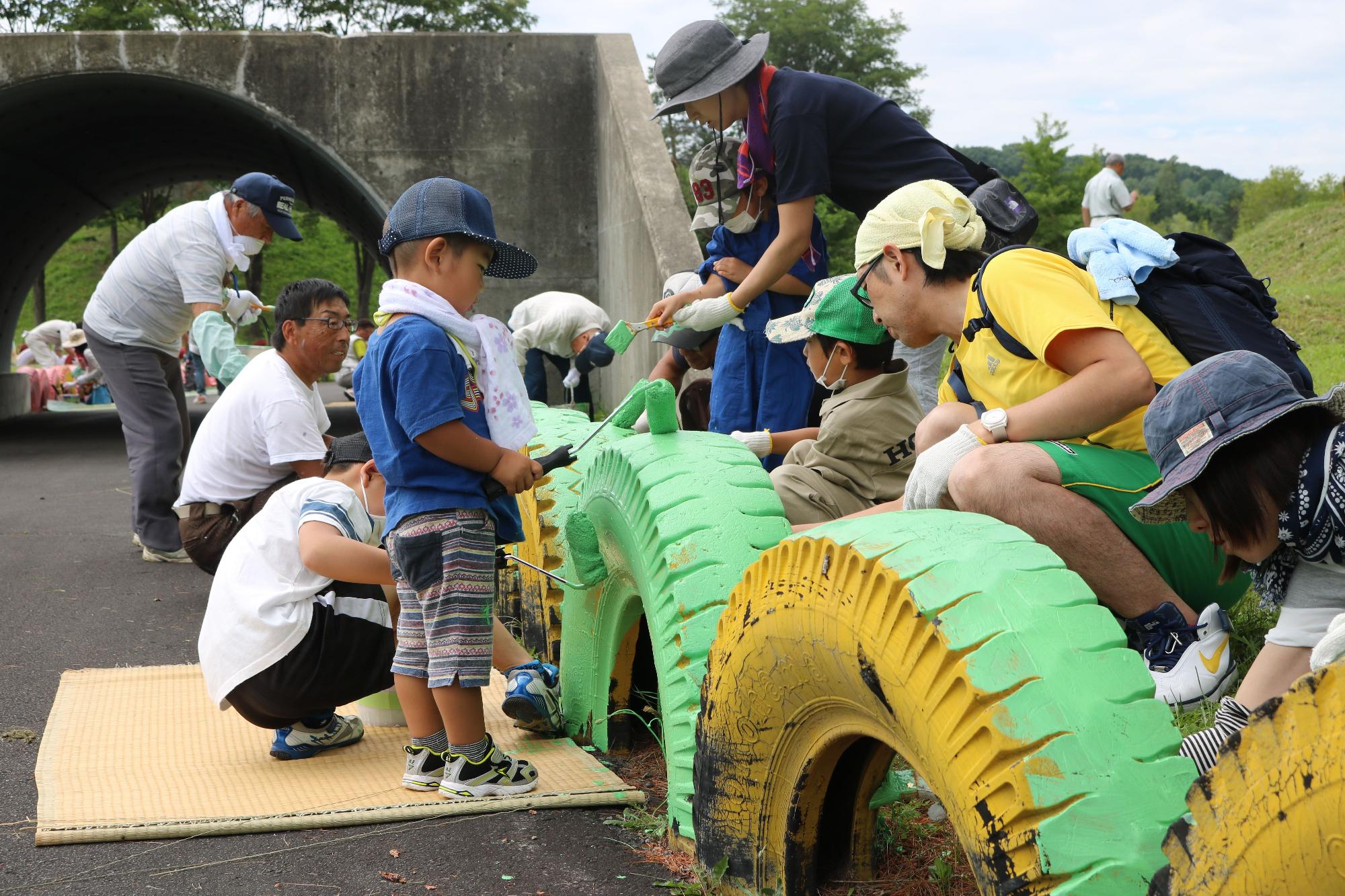 横一列に並んだタイヤの周りに子供や大人が集まり、黄色のタイヤを緑に色塗りをしている写真