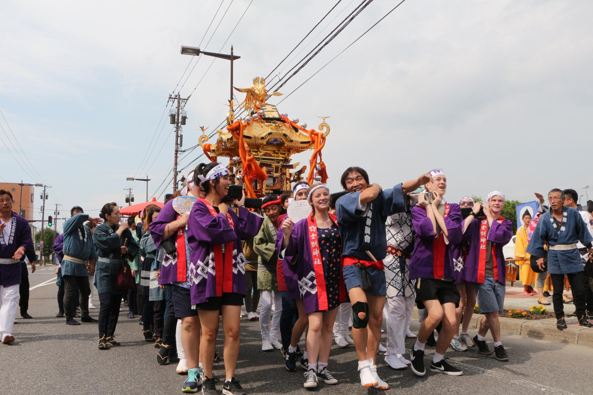 紫色の法被を着た人達が神輿を担ぎながら道路を歩いている写真