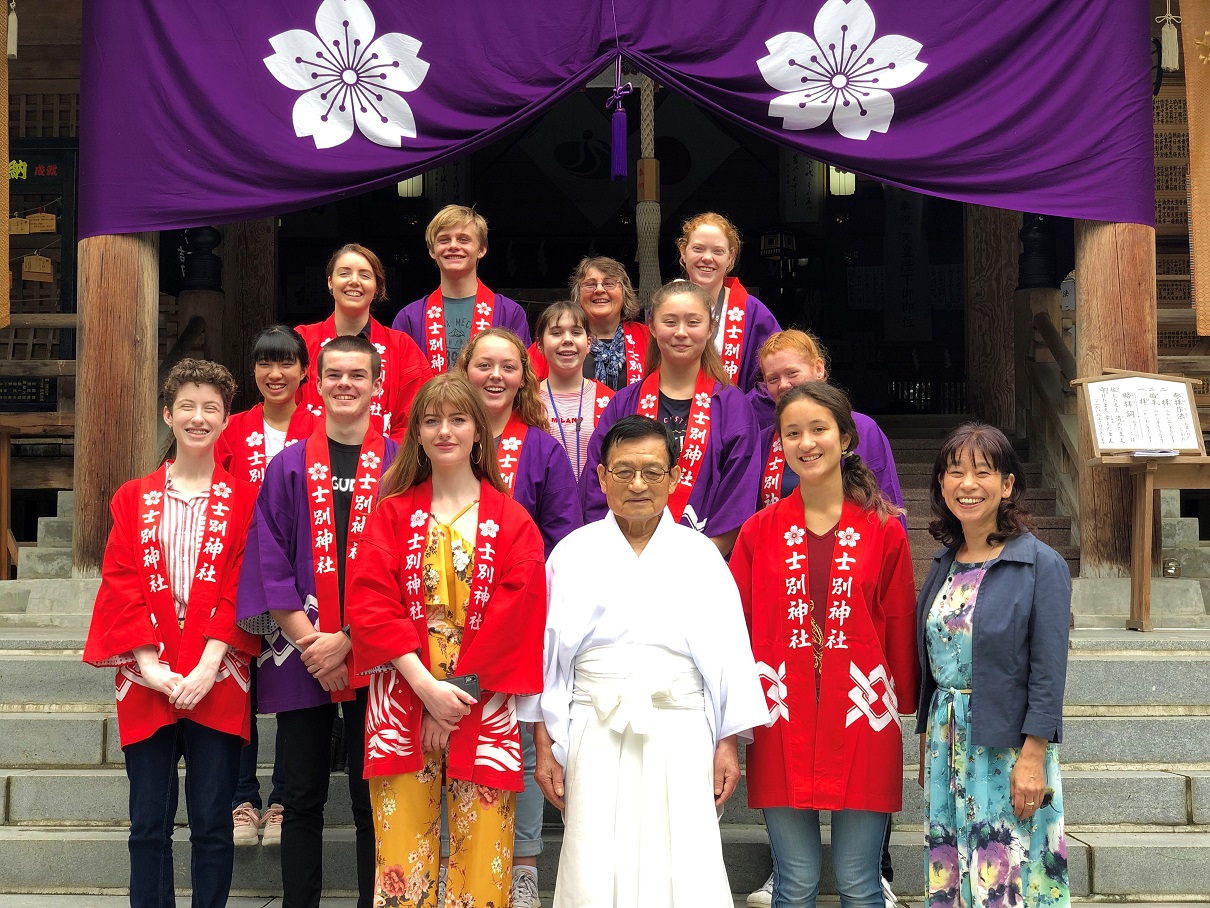 赤や紫の法被を着た留学生たちが神社の宮司さんと一緒に記念撮影をしている写真