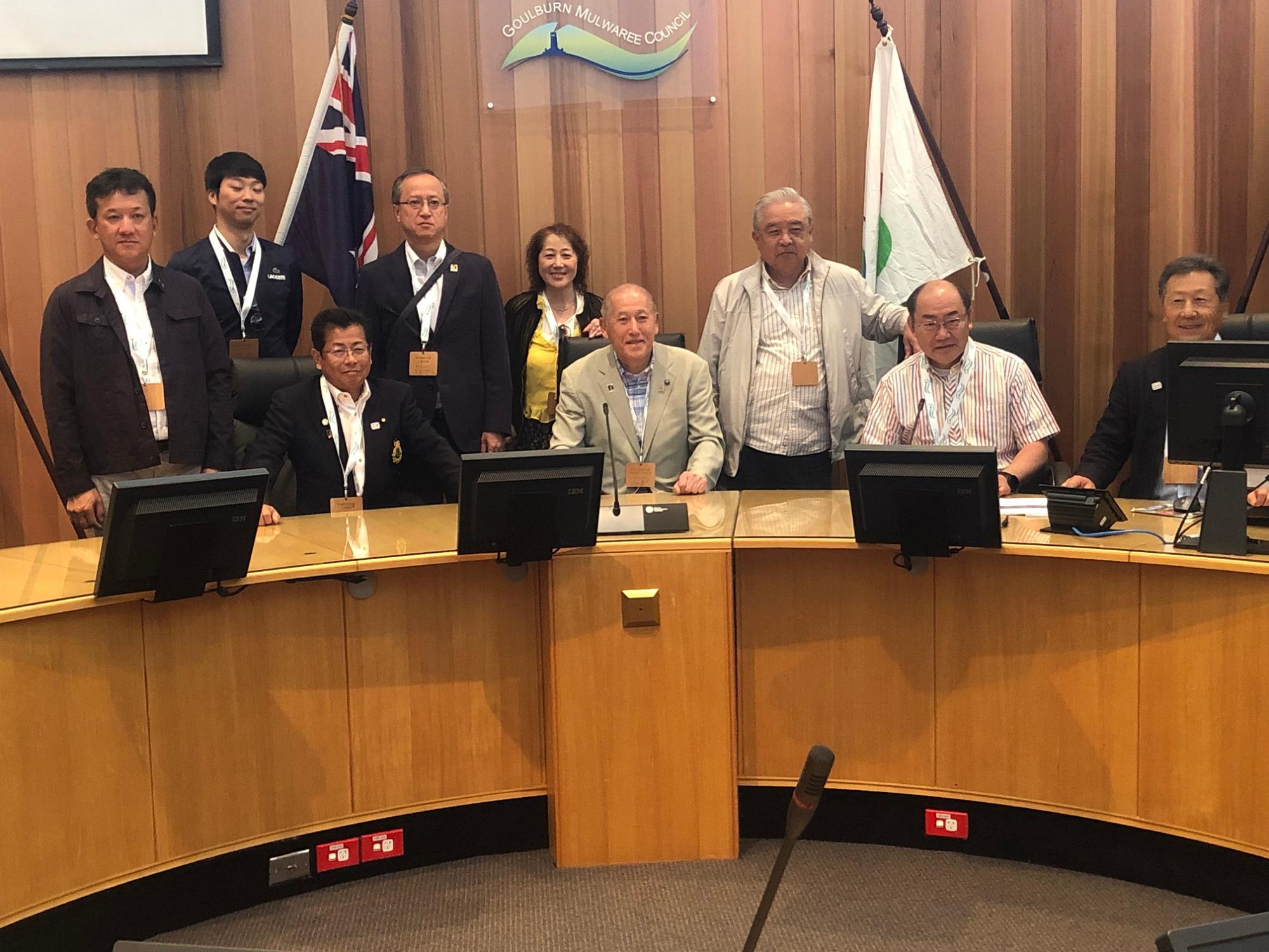 オーストラリアと士別市の旗の前の机で9名の参加者の人達が記念撮影をしている写真