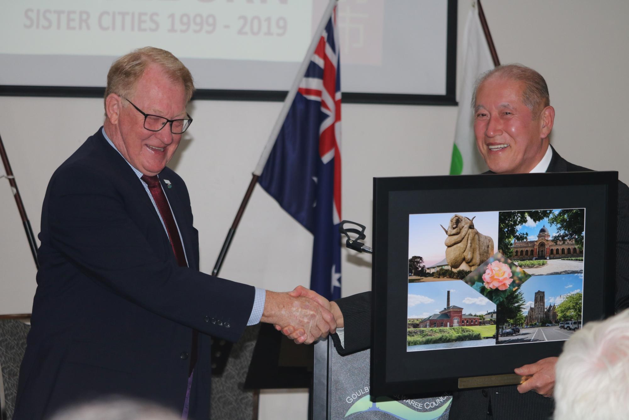 オーストラリアの国旗の前で男性2名が笑顔で握手をしている写真