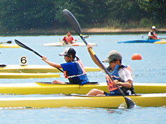 ゼッケンを付けた2人の選手がそれぞれ黄色いカヌーに乗りパドルで漕いでいるカヌー大会の写真