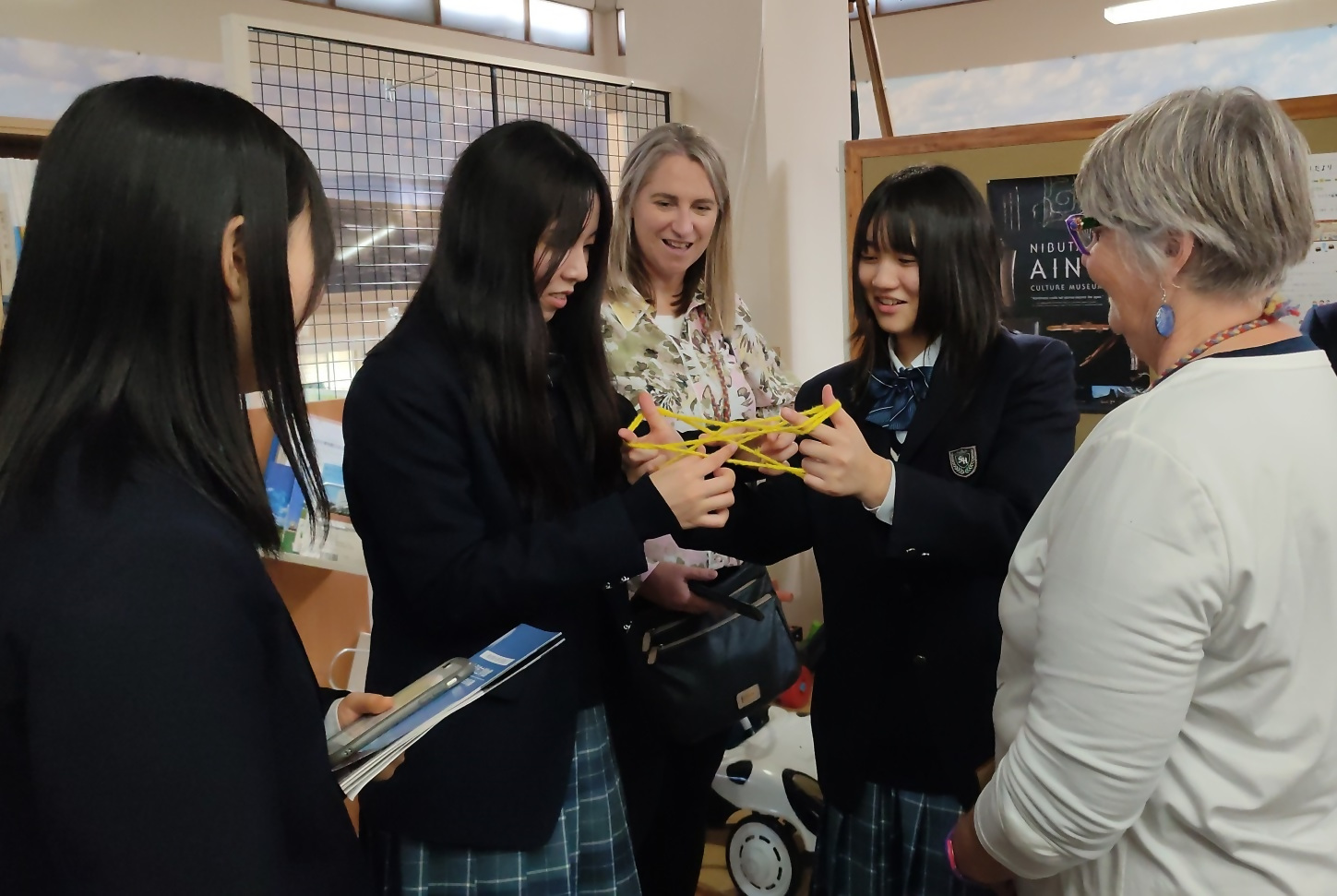 2人であやとりをする女子高生とその様子を見ている3名の女性の写真