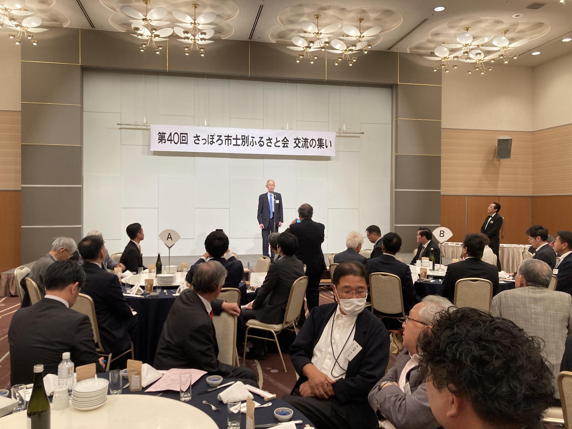 さっぽろ市士別ふるさと会交流の集いの横断幕の下の舞台で1人の男性の挨拶を会場の方々が見ている写真