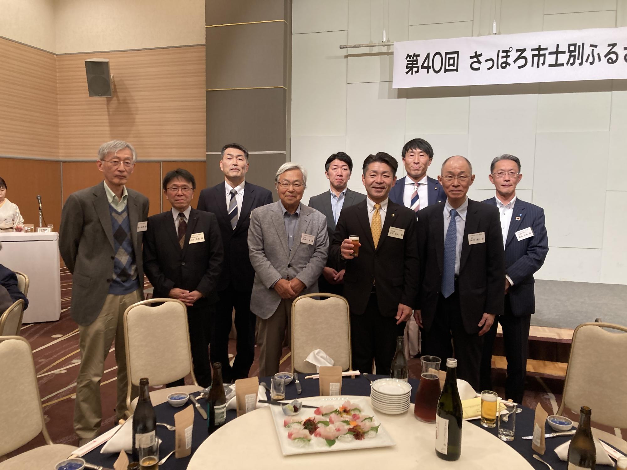 赤いテーブルクロスの丸いテーブルを囲んで右端にピンクの着物姿の女性が並び、男性7名女性5名、総勢12名の方が記念撮影をしている写真