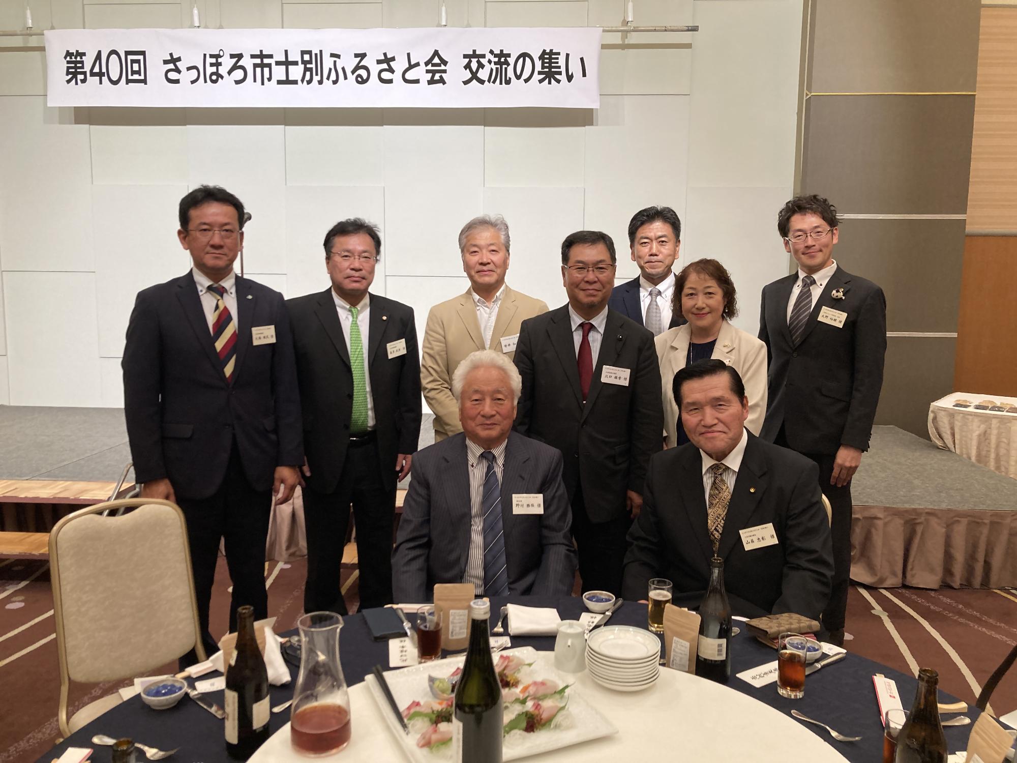 赤いテーブルクロスの丸いテーブルに料理や飲み物が並び、テーブルを囲んで右端にピンクの着物姿の女性がマイクを持ち並び、男性8名女性4名、総勢12名の方が記念撮影をしている写真