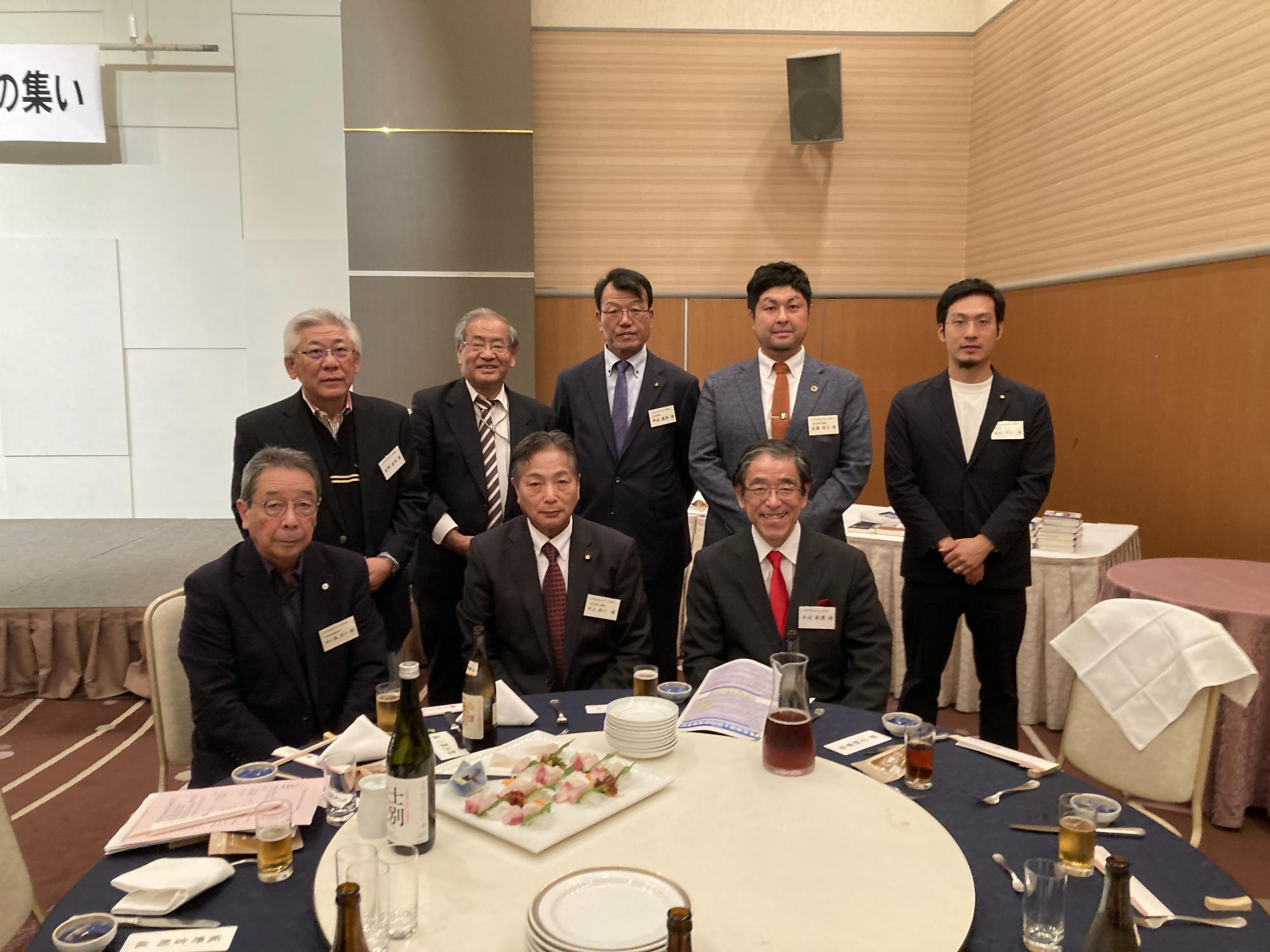 赤いテーブルクロスの丸いテーブルに料理や飲み物が並び、テーブルを囲んで中央にピンクの着物姿の女性が座り、男性11名、総勢12名の方が記念撮影をしている写真