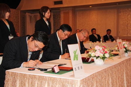 女性が2名が後ろに立っている、机で3名の男性が調印式で署名をしている写真
